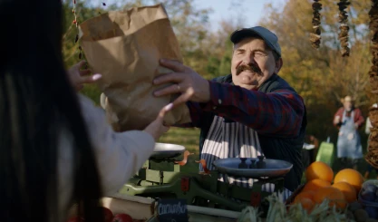Vendor at farmers market handing paper bag to customer, with produce and scale display